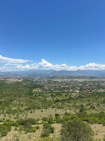 This is a landscape photograph taken from an elevated vantage point, likely a hill or mountain overlooking the city of Liješnje, Montenegro. The sky is a vibrant blue, with scattered white cumulus clouds. In the distance, a range of hazy blue mountains forms the horizon. Below the mountains, the city of Liješnje is visible, a sprawling urban area with a mix of modern buildings and smaller residential structures with red-tiled roofs. The foreground is dominated by a rugged, vegetated terrain with dry grass, scattered bushes, and small trees. The overall impression is a sunny day with a clear view of a city nestled within a mountainous landscape.