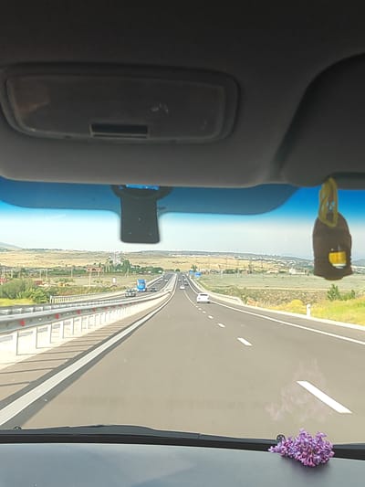 This media file captures a view from inside a vehicle traveling on a highway, likely in Oshakan, Armenia. The scene depicts a sunny day with a clear blue sky, suggesting warm weather. The highway stretches ahead, with multiple lanes marked by white lines. Several cars are visible on the road, indicating ongoing traffic. To the left of the highway, a guardrail separates it from a grassy embankment and then a landscape of rolling hills dotted with sparse vegetation and some scattered buildings, typical of a rural or semi-rural setting.
In the foreground, from the perspective of the vehicle's interior, a dashboard is visible, with a bouquet of purple flowers placed on it. Hanging from the rearview mirror is a brown car air freshener. The interior of the car suggests it is a passenger vehicle, with elements like the sun visor and the windshield providing the frame for the external view. The overall impression is one of a pleasant journey through a scenic, sunlit environment. There is no visible text in the media file.