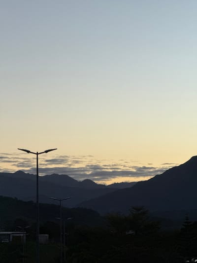 This vertical-format image depicts a twilight landscape in Dumbéa, France. The sky, transitioning from light blue overhead to a pale yellow-orange at the horizon, occupies the upper two-thirds. A band of horizontal, grey stratiform clouds is positioned above the distant mountain range. Layered, dark mountain silhouettes define the midground. In the foreground, three T-shaped streetlights with dual horizontal fixtures are silhouetted against the sky, the largest positioned in the left foreground. Dense, dark foliage occupies the lower portion of the frame, partially obscuring indistinct rectangular structures and a light-colored vehicle on the lower left. No individuals or active interactions are visible. The scene is characterized by low light, creating strong silhouettes across the foreground and midground elements.