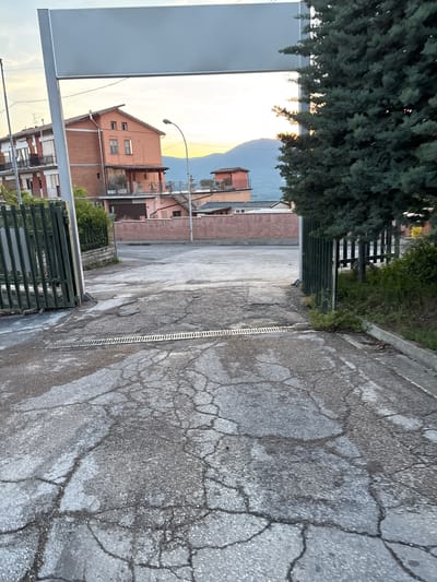 This image, set in Camerino, Italy, captures an outdoor scene, likely an alleyway or access road leading towards residential buildings and mountains. The perspective looks down a cracked asphalt path on the right side of the frame, which features a long, metallic drainage grate running alongside it. The asphalt shows signs of wear, including numerous cracks and some damp patches.

On the left side, a large, dense evergreen tree dominates the foreground, its dark green foliage contrasting with the bright sky behind it. Below the tree and further into the scene, a dark green metal fence or gate is visible.

In the mid-ground and background, several residential buildings with terracotta-colored facades and roofs are present, some appearing to be two to three stories tall with balconies and various window styles. A tall street light with a curved arm stands between the foreground elements and the buildings. In the distance, beneath a bright, clear sky, a range of soft blue mountains is visible. The sky exhibits a warm, golden hue near the horizon, suggesting late afternoon or early evening.

A large, empty grey sign or billboard frame stands prominently in the lower left portion of the image. No people or explicit activities are depicted. There is no visible text.