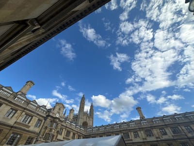 An upward-angle, low-light photograph captures the King's College Chapel in Cambridge, United Kingdom, under a bright blue sky dotted with fluffy white clouds. The chapel's iconic Perpendicular Gothic architecture, with its soaring twin towers and intricate stonework, dominates the center of the frame. Surrounding the chapel are the stone buildings of King's College, characterized by their ornate facades, numerous windows, and various architectural details like chimneys and decorative railings. The perspective is such that the buildings appear to lean into the frame from the left and right, creating a sense of depth. A white, tent-like structure is partially visible in the foreground, obscuring the very bottom edge of the image. The overall impression is one of grandeur and historical significance, set against a dynamic sky. There are no people or animals visible in the scene.