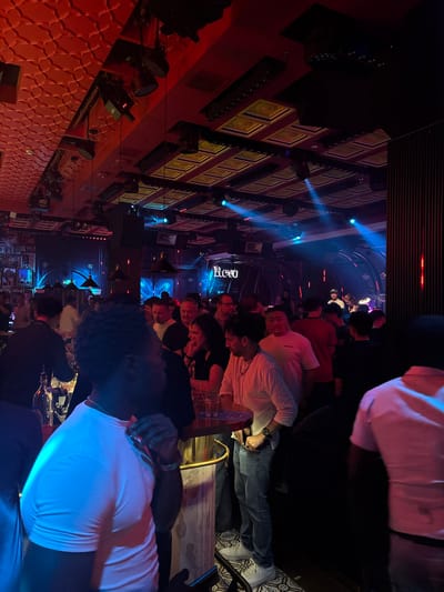 The image depicts a dimly lit, upscale nightclub or lounge scene in Jing'an District, China. A group of people are gathered, some standing and some sitting at tables, with a focus on socializing and enjoying the atmosphere. The environment is characterized by sophisticated decor, featuring ornate wall paneling with framed artwork and modern lighting fixtures. Blue and red spotlights create a dynamic and energetic ambiance, illuminating the space and highlighting the patrons.
The activity appears to be a social gathering, possibly a celebration or a night out. People are dressed in casual to semi-formal attire, suggesting a relaxed yet stylish event. The expressions on their faces are varied, but the overall mood seems to be one of enjoyment and engagement. Some individuals are looking at each other, suggesting conversation or interaction, while others are looking forward, perhaps at a performance or the general scene.
Notable details include the intricate patterns on the red wall, the various lighting equipment mounted on the ceiling and walls, and the presence of what appears to be a bar area in the background, hinted at by bottles and glasses. The time of day is likely evening or night, given the artificial lighting. There is no indication of weather conditions, as the scene is entirely indoors. The presence of the "REGO" text on a sign in the background provides a specific location cue, possibly a club name or a brand. The overall impression is one of a vibrant and lively nightlife venue.
