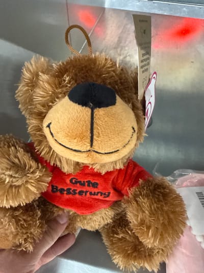 An overhead, medium shot shows a close-up of a brown teddy bear wearing a red shirt with the German words "Gute Besserung" (Get Well Soon) embroidered in black. The bear has a black nose and stitched eyes and mouth. A hand is visible holding the bear's left arm. The bear is hanging from a looped string attached to its head. In the background, a metallic surface reflects red light. A tag is attached to the bear's neck with text and a graphic. The image is not specific to Chociule, Poland.