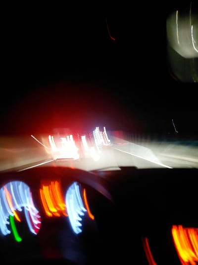 A blur of light streaks across a dark background. Predominantly red, orange, blue, green, and white, the colors swirl and merge. Some streaks resemble the distorted glow of car tail lights, while others appear as illuminated signage or streetlights. The effect is abstract and frenetic, suggestive of movement or speed. The image lacks a defined subject.