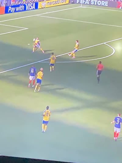 The image shows a soccer match in progress on a grass field with white line markings. Several players in yellow and blue uniforms are visible, as well as players in blue and white uniforms. One player in red is also present. The players are engaged in various actions typical of a soccer game, such as running and kicking the ball. In the background, a banner with the text "Pay with VISA" and "mercadolibre" is visible, suggesting a commercial sponsorship. The scene is outdoors, and the lighting indicates it could be daytime or under artificial lights. There is no information in the image to suggest the location is Zastavna, Ukraine.