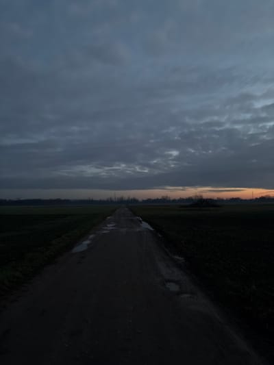 A long, straight dirt road stretches into the distance, flanked by dark fields. Puddles reflect the overcast sky. A band of orange light appears on the horizon. The sky is filled with dense, gray clouds. The left side of the image is significantly brighter than the right, which is dominated by deep shadows. The perspective emphasizes the road's vanishing point, creating a sense of depth and leading the eye toward the horizon.