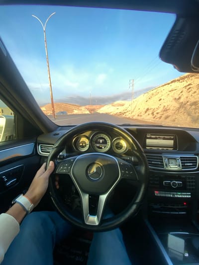 A person is driving a Mercedes-Benz on a highway. The driver's hand is visible on the steering wheel, wearing a watch. Outside, there is a clear blue sky and a mountainous landscape with power lines. The car's dashboard is visible, displaying information on the screen. The image is taken from the driver's perspective, capturing the interior of the car and the view through the windshield.
