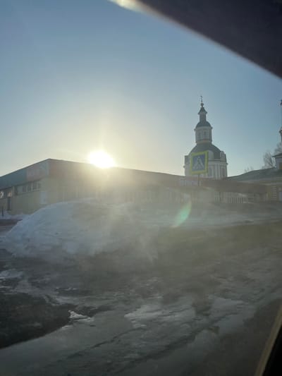 The image depicts an outdoor scene in Чутырь, Russia, under bright, low-angle sunlight, causing significant lens flare and glare. The foreground consists of dark, wet pavement and melting snow/slush, including a large, irregular snowdrift positioned left of center. In the midground, a single-story commercial building with light-colored walls and a red band displays Cyrillic signage, including "ПЯТЁРОЧКА" (Pyaterochka) and "МАГНИТ" (Magnit). Behind and to the right of this commercial building, a prominent Russian Orthodox church features a green dome, white facade with architectural details, and a golden cross atop its steeple. A yellow pedestrian crossing sign is visible on a pole in front of the church, partially obscured by the commercial building's roofline. The perspective suggests the image was captured from inside a vehicle. No individuals are visible.