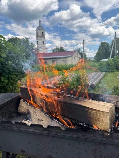The image shows a close-up of logs burning in a dark metal barbecue grill. Flames and smoke rise from the wood. In the background, a light-colored building with a red roof is visible, as well as a tall, slender tower with a dome. Green trees and vegetation surround the area, and a bright blue sky with white clouds is overhead. The scene appears to be set in a rural or semi-rural location. The specific location is the village of Luptyug, Arkhangelsk Oblast, Russia.