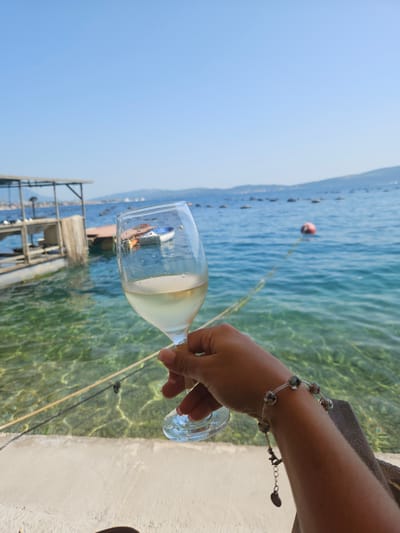 A hand holding a glass of white wine is shown in the foreground, with a bracelet on the wrist. In the background, a clear blue sea is visible, with a wooden structure and a few boats in the water. The sky is clear and blue. The location is Jošice, Montenegro.