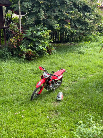 A red and white dirt bike is parked on a lush green lawn in Vang Vieng, Laos. A matching helmet rests on the grass next to the motorcycle. The background features dense green foliage and a portion of a wooden structure. The scene appears to be outdoors in a natural environment.
