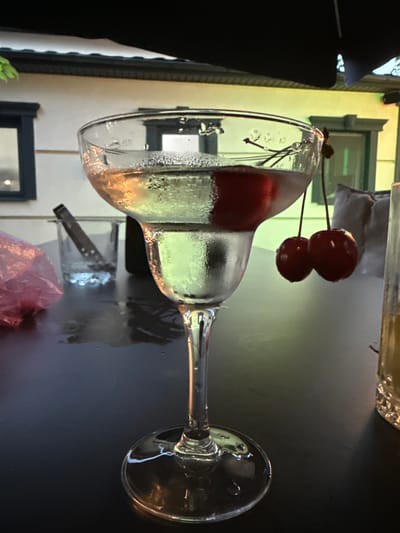 This image depicts a close-up of a margarita glass filled with a light-colored beverage, possibly a cocktail, with condensation on the outside of the glass. Two red cherries are suspended from a stem above the drink, with one cherry partially submerged in the liquid. The glass rests on a dark, reflective surface, likely a table. In the background, out of focus, a building with white siding, dark window frames, and a decorative facade is visible. The overall lighting suggests an evening setting, possibly outdoors or in a dimly lit establishment. The location is specified as Edineț, Moldova, although this context is not visually evident within the image itself.