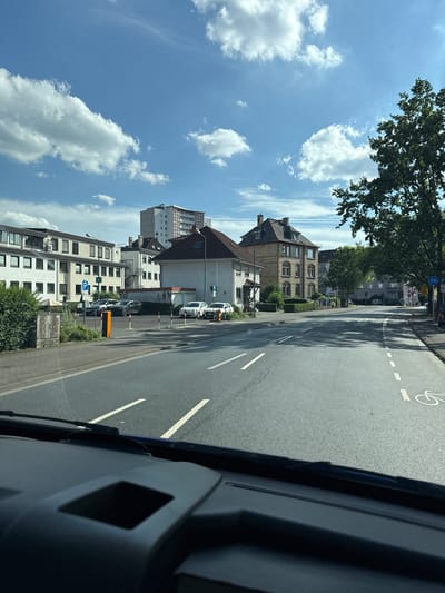 This is a photograph taken from inside a vehicle, looking out at a street scene in Giessen, Germany. The perspective is from the driver's seat, showing the dashboard in the foreground. The street is paved and has lane markings. On the left side of the street, there is a sidewalk, a building with many windows, and a parking lot with several cars and parking signs. Further down the street, there are more buildings, including a taller apartment building and older, multi-story houses with pitched roofs. On the right side of the street, there are trees and more buildings, as well as a bicycle lane symbol painted on the road and a traffic sign. The sky is blue with white, puffy clouds, indicating a clear day. The overall impression is of a typical urban or suburban street in Germany on a sunny day.