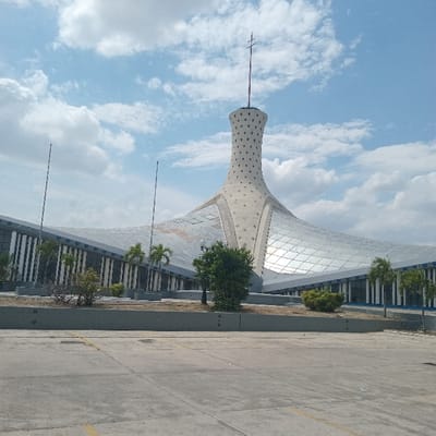 The image displays an exterior, eye-level view of the Manto de María (Mantle of Mary) monument in Barquisimeto, Venezuela, under a partly cloudy blue sky.

The central subject is a monumental, modern architectural structure. It features a tall, slender, conical main tower, light-colored and textured with numerous small circular indentations, tapering upwards to a spire crowned with a Christian cross. An expansive, undulating roof system, composed of a grid of reflective or translucent panels, extends outward from the tower's base. Below this canopy, the building's lower structure presents a facade with vertical striped elements in dark and light hues.

In the foreground, a wide, grey concrete pavement with visible expansion joints and faded yellow markings occupies the lower portion of the frame, indicating a large parking area or plaza. This paved area transitions to a raised concrete planter wall separating it from the monument. Within the planter and directly adjacent to the building, various types of vegetation are present: multiple green shrubs, several small bushy trees, and distinct slender palm trees positioned along the building's perimeter. To the left, two tall, dark, slender poles stand vertically. The scene is brightly illuminated, indicating daylight. No individuals are present.