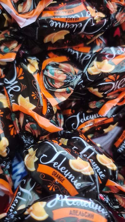 The image displays a close-up, overhead view of a pile of individually wrapped candies. The candies are predominantly black with orange and white accents. Each wrapper features stylized lettering that appears to spell "Jeleynie" in white, cursive script, and the word "ORANGE" in smaller, white, uppercase letters on an orange band. Sliced oranges are depicted as part of the wrapper design, along with decorative floral elements.

Visible text on the wrappers includes "Jeleynie", "ORANGE", and what appears to be "АПЕЛЬСИН" in Cyrillic, which translates to "APELSIN" or "ORANGE". Other Cyrillic text is present but is partially obscured or too blurry to be definitively read. The overall impression is of a sweet treat, likely with an orange flavor, packaged for sale. The lighting suggests an indoor setting, possibly a store or a home, as there are no strong shadows or indicators of natural light. The focus is entirely on the candies themselves, creating a sense of abundance or a collection of many. There are no people or animals visible in the frame. The setting is generic, focusing on the product.