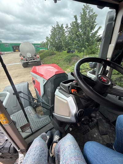 A view from inside the cab of a Massey Ferguson 8S.425 tractor, showing the interior elements: steering wheel with the Massey Ferguson logo, dashboard, floor pedals, and seat. Outside the cab, a green building is visible on the left, along with a large water or liquid tank trailer. The background includes green grass, trees, and a cloudy sky. The photo was likely taken in an agricultural or industrial setting. The location of Gorna Mitropolia, Bulgaria is implied.