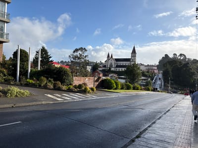 The image shows a street scene in Puerto Varas, Chile, with a view of the Sagrado Corazón de Jesús Church in the background. The foreground features a wet asphalt road with a crosswalk. On the left side of the frame is a section of a building with balconies, and some greenery. On the right, a sidewalk with patterned pavers is partially visible. Several pedestrians are walking on the right side. Vehicles can be seen driving down the street towards the church. The sky is blue with scattered clouds.