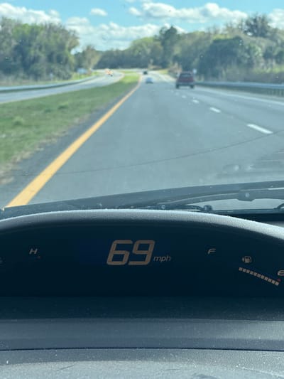The image presents a first-person view from inside a vehicle traveling on a multi-lane highway in Gainesville, United States, under a partly cloudy sky. The vehicle's digital speedometer on the dashboard displays "69 mph," and the fuel gauge shows approximately three-quarters full. The dashboard surface appears dusty, with windshield wiper blades visible at the base of the windshield. Ahead, the asphalt highway extends, featuring a solid yellow line to the left of the current lane, separating it from a grassy median. Further left, another highway lane is visible, followed by a treeline. To the right, a second lane is present, with white dashed lane markings visible beyond. Several vehicles are discernible in the distance, including a red SUV-like vehicle in the right lane and a lighter-colored vehicle further ahead in the left lane. The road appears to curve gently to the right in the far distance, bordered by dense treelines on both sides. No specific individuals are visible outside the vehicle.