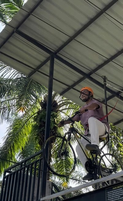 A man wearing an orange helmet and glasses is riding a bicycle on a raised platform. He is wearing a purple short-sleeved shirt and white pants. The bicycle is black and yellow. A safety harness is attached to him and a cable. Behind him, palm trees are visible against a bright sky. A metal roof structure is overhead, supported by dark beams. Below the platform, a black metal railing is visible. A sign with "PARK INSTRUCTIONS" is partially visible at the bottom right. This scene is situated at the Bintang Meriah adventure park in Bintang Meriah, Indonesia.