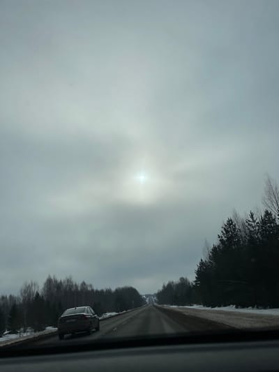 The scene depicts a rural road in Bol'shoi Talmaz, Russia, viewed from inside a moving vehicle. A two-lane asphalt road, appearing damp, stretches into the distance under an overcast, grey sky. A bright, diffuse light source, consistent with the sun obscured by clouds, is visible in the upper central portion of the sky. Snow and slush line the road shoulders. Dense coniferous trees border the right side of the road, while bare deciduous trees are visible on the left and in the background. A dark-colored sedan, bearing a Russian license plate, travels ahead in the right lane, receding from the viewer. The lower edge of the image shows the dark top of a vehicle's dashboard. No individuals are discernible.