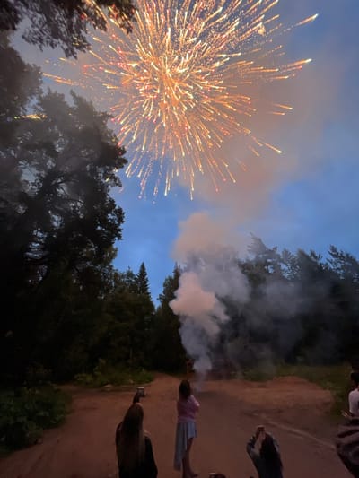 In Simagino, Russia, at dusk, a group of individuals observe a fireworks display in a wooded area. The fireworks explode in the sky, creating a brilliant burst of orange and yellow sparks, accompanied by plumes of smoke. The foreground is dominated by a dirt path, with several people silhouetted against the twilight sky. One person in the foreground is holding up a phone, presumably to record the event. Tall trees frame the scene on either side, their dark silhouettes contrasting with the bright sky. The overall atmosphere is one of shared enjoyment and spectacle, set against a natural, outdoor backdrop.
