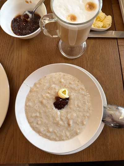A top-down view shows a breakfast scene on a wooden table, possibly indoors. A bowl of porridge sits on a white plate, topped with a curl of butter and a small dollop of dark jam or compote. A spoon rests on the edge of the plate. To the left is a glass mug filled with a light brown beverage, possibly a latte, topped with foamed milk. Next to the mug, there's a small dish containing more of the dark jam or compote with a spoon in it. A small white bowl with curls of butter is visible near a knife. A spoon rests on the edge of the plate containing the porridge. The location is most likely a private residence or cafe in Тэльмінскі сельскі Савет, Belarus.