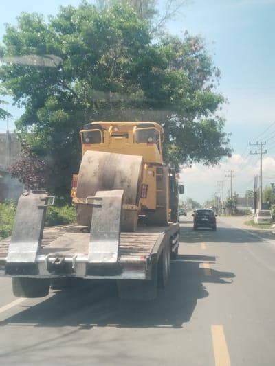 A yellow construction steamroller is transported on a flatbed truck along a road marked with yellow lines. The scene is in Blang Lilue, Indonesia, marked by utility poles along the roadside. A dark SUV follows behind the truck, and a white van is parked further down the road. Trees are visible in the background.
