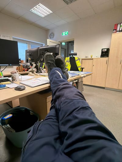 An individual's legs, clad in dark blue work trousers with a white diagonal stripe on the outer right leg, and dark lace-up work boots, are depicted resting on a light wood-grain office desk. The perspective is first-person, looking down at the legs and across the office environment.
On the desk surface, directly behind the boots, are two black computer monitors on a shared stand. To the left of the boots, a brown wooden desk organizer holds pens, a white mug with an illustration, a dark gray computer mouse, and various documents, including one with a yellow sheet. Cables are visible on the desk. A dark gray or teal waste bin containing papers and a small object is on the floor to the left of the legs.
In the midground, behind the desk, a light-colored office chair with a black seat and backrest features a bright yellow high-visibility safety vest draped over its back. To the right, a light wood-grain storage cabinet supports a black coffee machine, a yellow cup, and a clear container. Further right, a taller, matching cabinet holds red and green binders on an upper shelf.
The background shows light-colored walls. A window with light-colored horizontal blinds is on the left. A doorway leads to another area, with an illuminated green exit sign above it and a round wall clock to its left. The ceiling comprises square acoustic tiles with a centrally located, illuminated rectangular fluorescent light fixture. The scene is located in an office within Grevenmacher, Luxembourg.