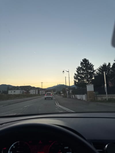 The image depicts a roadway scene at dawn or dusk in Kaysersberg Vignoble, France, captured from within a vehicle. The foreground reveals the top section of a dark steering wheel and dashboard, with an instrument cluster showing red digital readouts including "220" and "260". The main subject is a two-lane asphalt road extending into the distance, marked by a solid white line in the foreground transitioning to a dashed white line further ahead. A white SUV or hatchback, possibly a service vehicle due to a red and white chevron pattern on its rear, is visible ahead on the road with its rear lights illuminated.

To the left of the road, several residential buildings with light-colored facades and pitched roofs are visible, backed by distant, undulating hills. To the right, a sidewalk or paved shoulder borders a property enclosed by a low white wall with distinct pillars, behind which dense, dark trees are present. A black streetlamp with an ornate light fixture stands prominently on the right. Utility poles with overhead wires line both sides of the road. The sky exhibits a gradient from pale blue at the top to a warm yellow-orange closer to the horizon, and a faint aircraft contrail is visible in the upper left sky. No individuals are discernible in the scene.