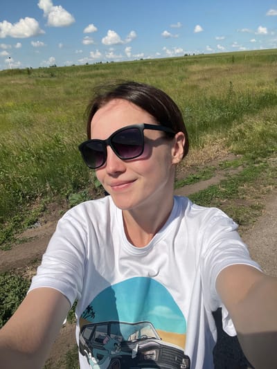 A woman is taking a selfie outdoors in a field under a blue sky with white clouds. She is wearing sunglasses and a white t-shirt with a graphic of a car and cartoon characters. The background shows a grassy field with a dirt path. The location is the city of Mikhailovka, Russia.