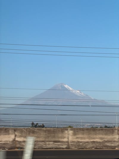 The image presents an outdoor scene likely observed from a moving vehicle in or near Santa María Zacatepec, Mexico. The central background is dominated by a large, snow-capped stratovolcano (Popocatépetl), its peak covered in white snow against a clear, light blue sky. Atmospheric haze partially obscures the mountain's lower slopes and the distant landscape. A horizontal band in the mid-ground reveals distant urban or suburban development, characterized by scattered structures and green tree cover. Several tall, slender streetlights or utility poles are visible in this mid-ground area. The immediate foreground features a concrete barrier, weathered and textured, running horizontally across the lower portion of the frame. Below this barrier, a dark road surface is visible, blurred due to motion. Multiple dark, horizontal utility lines traverse the entire image width, layered over the sky, mountain, and mid-ground elements. A single, light-colored, vertically oriented object, heavily blurred, is partially visible in the lower-left foreground, consistent with an object passed at speed. No individuals are depicted.