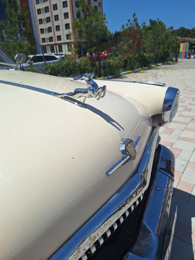 An exterior shot captures the front section of a vintage, cream-colored automobile, possibly parked outdoors in an urban setting. The hood is adorned with a chrome hood ornament depicting a leaping gazelle. A chrome trim piece runs along the hood's edge, and a distinctive emblem is mounted on the front fender. The car's front headlight, encased in chrome, is visible on the right. The vehicle's chrome bumper, exhibiting some signs of wear and rust, is prominent in the foreground, and the grille is partially visible beneath it.
The background reveals a tiled pedestrian area. Beyond this, a modern, multi-story building with numerous windows stands out. Green trees are interspersed throughout the scene, and a bright blue sky dominates the upper portion. In the far distance, figures of people and other vehicles can be vaguely discerned. The presence of a colorful art installation, featuring a grid of colored squares, adds a touch of vibrancy to the background. The lighting suggests a sunny day. The context of the city of Khudat, Azerbaijan, is provided, indicating the geographical setting of this scene.