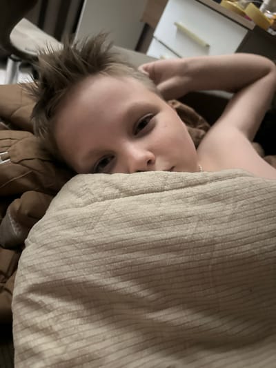 A young boy with spiked brown hair rests on a bed, partially covered by a tan blanket. His right arm is bent behind his head, and his left eye is visible as he looks at the camera. The background suggests an indoor setting, possibly a bedroom, with hints of furniture and a dresser with items on top. The image is a close-up, portrait-style shot.