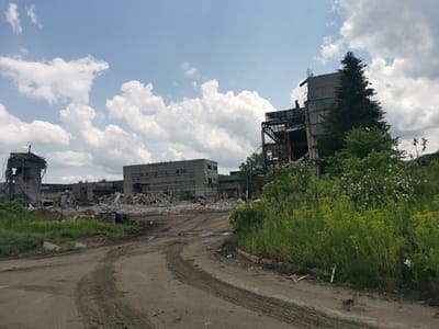 This image captures a desolate, overgrown industrial area under a partially cloudy sky. The scene is dominated by a collection of large, derelict concrete buildings in various states of decay, suggesting abandonment or demolition.
In the foreground, a wide, unpaved dirt road with visible tire tracks curves through the landscape. Patches of green grass and yellow flowering weeds line the road, particularly prominent on the right side, where lush vegetation is beginning to reclaim the space.
The midground features extensive piles of rubble and debris, remnants of collapsed structures. Several multi-story industrial buildings, constructed from concrete, stand beyond these piles. Many windows are broken or missing, and structural elements like metal beams are exposed. To the far left, a round or cylindrical structure is visible, partially collapsed. On the right side, a tall, dark green conifer tree stands prominently next to and partially obscures one of the skeletal industrial buildings, which has exposed metal framework. Behind some bushes on the right, a "HYUNDAI" excavator is partially visible, hinting at ongoing or recent demolition work.
The sky above is bright and clear in parts, with numerous fluffy white cumulus clouds scattered across a light blue expanse. The lighting suggests it is daytime, possibly late morning or early afternoon, with decent visibility. The overall impression is one of decay and nature slowly reclaiming an abandoned human-made environment in Zhivkovo, Bulgaria. No people are visible.