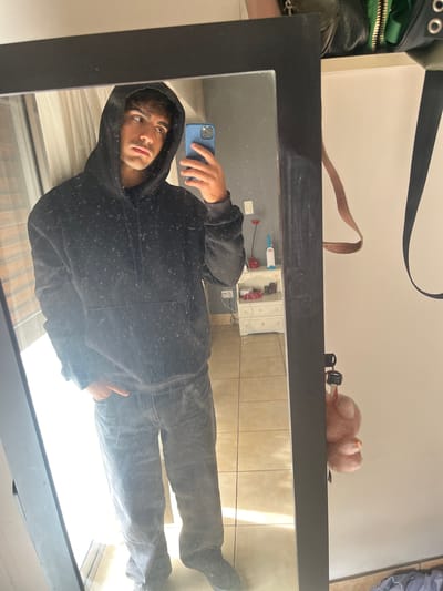 The image displays an indoor scene, rotated 90 degrees counter-clockwise from its natural orientation, showing a person taking a mirror selfie.

At the center is a large, black-framed mirror, reflecting a young man. He has dark hair and a serious or contemplative expression, looking slightly upwards. He is wearing a black hooded sweatshirt with the hood up and dark-colored pants. His right hand is extended, holding a blue smartphone (possibly an iPhone) towards the mirror to capture the selfie. The mirror surface shows numerous small specks, suggesting dust or minor smudges.

Behind the man's reflection, the interior of a room is visible, featuring a light-colored tiled floor and a white cabinet with two doors, adorned with small pink floral decals. Several objects, including what appear to be cleaning supplies (a red object, a white bottle, a blue brush), are placed on or near the cabinet.

On the real-world surface directly above the mirror's frame (appearing to the left of the image when rotated correctly), various items are arranged against a plain white wall. These include a black bag with green zippers and silver eyelets on its strap, a brown leather strap, a small, pink, fluffy animal figurine (possibly a pig) lying on its back, and a set of keys with a black fob.

The lighting suggests it is daytime. The overall setting appears to be a casual home environment in Sarandí, Argentina. No discernible text is visible in the image.