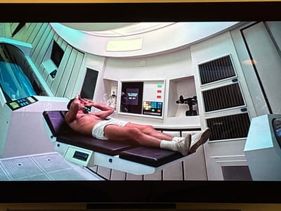 The depicted environment is a futuristic or laboratory interior, inconsistent with a typical location in Cabo Velas, Costa Rica. An adult male is positioned supine on a dark-padded examination table with a white, angular base. He wears white briefs, white socks, and white athletic shoes. His hands are placed over his face, covering his eyes and forehead. The room features smooth, curved white walls and angular structural elements. To the left, a prominent, large, translucent blue-green chamber with internal components and an integrated control panel displaying illuminated buttons is visible. To the right of the male, a central control panel features a screen displaying "00 01" and partially visible text, likely "THE WORLD," surrounded by various buttons and indicators. Above and to the right of this central panel, three vertically stacked rectangular units with dark, striped grilles are present. A black, multi-jointed mechanical device rests on a shelf near the central console. The environment is illuminated by bright, cool-toned white and blue-green light, creating a sterile and technologically advanced aesthetic.