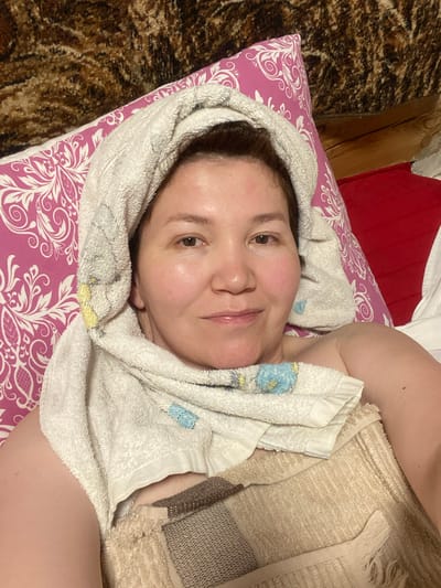 A woman lies on her back, wrapped in towels. A pink and white floral patterned pillow is beneath her head, and a brown, textured blanket is behind it. A red surface is visible to the right of the pillow. The woman's head is partially covered by a white towel, and another towel is draped across her chest. Her skin appears somewhat flushed, and there is a red mark on her forehead. The setting appears to be indoors.