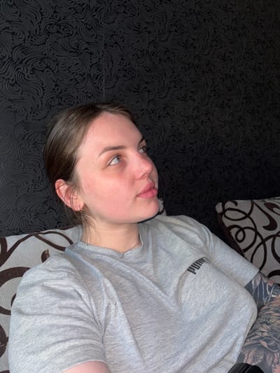 The image displays a young person, likely a woman, lying down indoors, viewed from an angled perspective. Her head is positioned towards the left of the frame, with her body extending to the right. She has fair skin, light eyes, and freckles visible on her nose and cheeks. She wears a small hoop nose ring and a hoop earring in her visible ear. Her lips are slightly parted, and her dark hair is brushed away from her face.

She is dressed in a light gray short-sleeved t-shirt, featuring the black "PUMA" logo on the upper chest/shoulder area. Extensive dark tattoos are visible on her right arm and shoulder. She appears to be resting on a dark surface with a subtle, embossed swirling pattern, possibly a bedspread or floor covering. A section of a lighter-colored cushion or pillow, adorned with a bold dark brown swirling pattern, is visible behind her head and beneath her right arm. The setting suggests a casual indoor environment, potentially a home in Chornomorskoe, Ukraine. Her gaze is directed upwards and to the side, conveying a contemplative or relaxed expression.