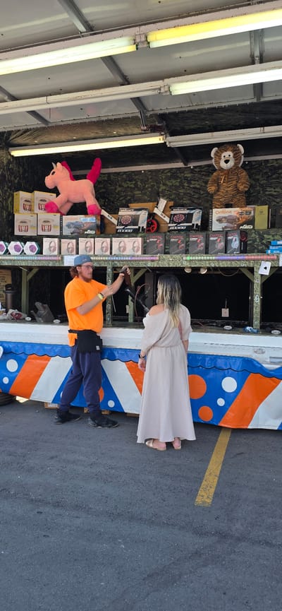 In Pickering, Canada, at what appears to be a carnival or fairground booth, a man wearing an orange t-shirt and dark pants is demonstrating a rifle to a woman. The woman is dressed in a light-colored, off-the-shoulder dress and sandals. Behind them, on shelves, are various prizes including stuffed animals (a pink unicorn and a tiger), boxes of headphones, and what seem to be dartboards. The booth itself is decorated with a colorful banner featuring orange and blue circles. The ground is asphalt, with a yellow line visible.