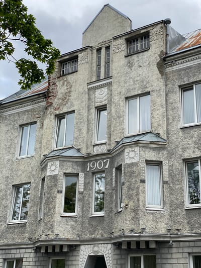 The image shows a close-up, eye-level view of the facade of a historic building in Sortavala, Russia. The building's exterior is a rough, textured grey stucco, showing signs of age and some weathering, with exposed brick in one section. The architectural style appears to be Art Nouveau or a related early 20th-century style, characterized by decorative elements such as floral motifs and molded ornamentation.
Prominently displayed on the central bay of the facade are the white numerals "1907", indicating the year of construction. Numerous windows are visible across different floors, mostly rectangular with white frames and blinds or curtains. Some windows are reflecting the overcast sky and surrounding trees.
The roofline is visible at the top, with some sections appearing to be made of corrugated metal that shows rust. There is a drainpipe running down the side of the building. At the bottom of the image, a portion of the building's base is visible, constructed with grey brickwork and featuring decorative circular motifs. The overall impression is of an aged, architecturally interesting building in an urban setting, with no people or active events depicted.