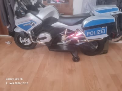 A gray and black toy BMW police motorcycle, labeled "POLIZEI," is indoors on a light-colored wooden floor. The motorcycle has a small support wheel extending from its base. The foreground is mostly empty, showing the wooden floor. Background elements include a gray garment, a wooden doorframe, and indistinct objects possibly covered by a white sheet or curtain. The image's lower-left corner displays the text "Galaxy S20 FE 1. Juni 2026 13:12." There is no evidence of a specific location within Bingen am Rhein, Germany.