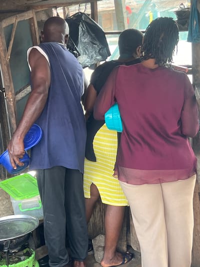 In Ijegun, Nigeria, the image shows a wooden structure, possibly a shop or stall, with several people inside. One individual is wearing a dark purple, semi-sheer blouse and light tan pants. Another person is clad in a yellow and white striped garment. A third person wears a black shirt and has dreadlocks. A bald man is dressed in a blue shirt and black pants. The individuals are holding small plastic containers. Additional objects in the scene include black plastic bags, metal objects, and green plastic containers. The overall impression is of a cramped, indoor space with a mix of people and belongings.