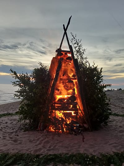 A large bonfire is burning on a sandy beach near the water's edge. The bonfire is constructed from logs and branches, creating a towering structure engulfed in flames. The fire is bright orange and yellow, casting a warm glow on the surrounding logs and the foliage that encroaches upon it. The sky above is a mix of gray and pale blue, with wispy clouds suggesting a late afternoon or early evening setting. The sun appears to be low on the horizon, as indicated by the soft, diffused light.

The beach is sandy, with footprints visible in the sand, indicating recent activity. There is some vegetation growing on the dunes behind the bonfire, with green leaves and some white flowers. The water of the sea is calm, with a gentle ripple reflecting the colors of the sky. In the distance, across the water, there appears to be a faint silhouette of land or structures. The overall mood is one of warmth and possibly celebration, with the bonfire as the central focus. There is no visible text in the image. The scene is set in Vārzas, Latvia.