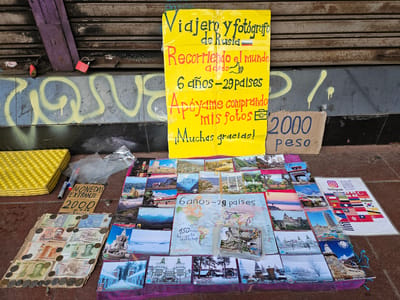 The image shows a street vendor's display in Osorno, Chile. The vendor, presumably a traveler and photographer from Russia, is selling postcards featuring scenes from around the world. A yellow handwritten sign advertises the vendor as a "Viajero y fotógrafo de Rusia" and states that they have been traveling the world for six years, visiting 29 countries. The sign asks for support by purchasing their photos and mentions "muchas gracias" (thank you very much). A cardboard sign indicates a price of "2000 peso" (Chilean currency).
The vendor's wares include a collection of postcards laid out on a cloth. A world map, marked with travel routes and handwritten text like "6 años-29 países" and "150 mil km haciendo autostop" (150,000 km hitchhiking), is displayed among the postcards. Foreign currency and coins are arranged on cardboard next to the postcards. There's also a small sign with an Instagram logo and a QR code. A selection of flags from different countries, presumably representing the vendor's travel destinations, is also shown.