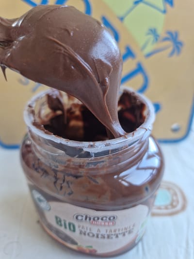 A close-up shot focuses on a spoon lifting creamy chocolate hazelnut spread from an open jar. The jar, labeled "BIO PÂTE À TARTINER NOISETTE," is partially filled with the spread. The background shows a yellow surface with blurred blue palm tree graphics, hinting at a possible picnic mat or beach towel. The scene appears to be indoors, on a white surface, likely a table. There is no discernible urban environment or any indication of the city of Fresnes, France.
