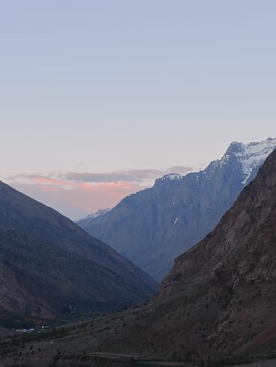 The image depicts a mountainous landscape, possibly during twilight or dawn, given the soft lighting and pastel colors in the sky. In the foreground, two large mountain slopes converge, creating a valley that recedes into the distance. The left slope is darker and appears to be covered in sparse vegetation. The right slope is a reddish-brown, rocky mountainside with scattered green shrubs.
Further in the distance, beyond the initial slopes, are more rugged mountains. Some of the higher peaks are capped with snow, reflecting the dim light. A soft band of pink and orange clouds stretches across the sky horizontally, just above the distant mountains.
In the valley floor, visible in the lower part of the image, there appears to be a small settlement. Several buildings are clustered together, and some lights are visible, suggesting human habitation. The area around the settlement is dotted with trees and appears to be more cultivated than the surrounding slopes. The overall impression is one of a remote, natural environment with a small human presence nestled within. The location is identified as Gemur, India.