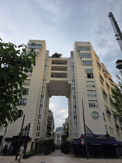 The image shows a street scene in Montrouge, France, with two modern apartment buildings flanking a walkway. The buildings have light-colored facades with numerous windows and balconies. A bridge-like structure connects the two buildings at an upper level. Trees and greenery are visible, including a large tree in the foreground partially obscuring the lower portion of the buildings.
On the ground level of one of the buildings, there is a storefront with signage indicating "Bar Le Mekc" and "Restaurant." Another storefront has signage for "FLEURS" (flowers) and mentions "Naissance-Mariage-Devil" (births-marriages-deaths) and "Ouvert 7 jours/7" (open 7 days a week). A person is walking on the sidewalk in front of the restaurant.
The sky is overcast, suggesting a cloudy day. The time of day is difficult to determine precisely but appears to be daylight. The architectural style of the buildings is contemporary. A traffic sign with red and blue circles is visible. The overall impression is of an urban street in a residential or mixed-use area.