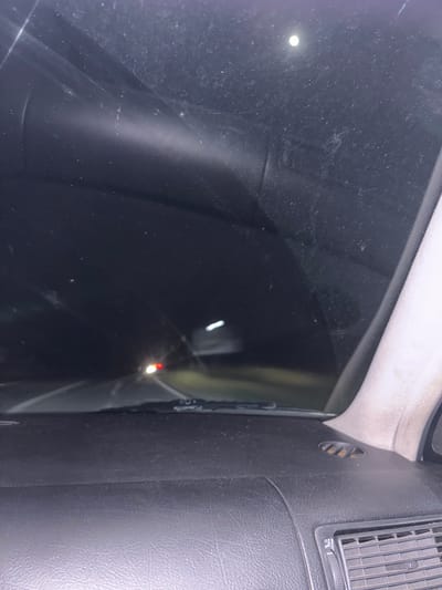 The image shows an interior view through a car window at night. The location is inferred to be Novo Leski, Bulgaria, based on the prompt context. The view is obscured by a dirty car window. A faint light source is visible through the darkness. A dashboard vent is visible on the right. The dashboard appears to be black plastic or leather. The headliner trim is visible in the upper section of the image. No people, actions, or interactions are visible. The environment is dark, with only reflected lights visible.
