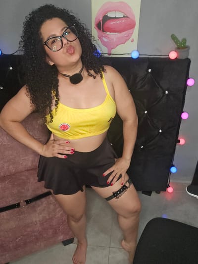 In a dimly lit indoor setting, possibly within Perímetro Urbano Cúcuta, Colombia, a woman with curly dark hair and glasses poses with her lips pursed. She is wearing a yellow sports bra and a black skirt with a garter. The background features a black tufted headboard adorned with lights, a pink sofa, and a poster of lips on the wall.