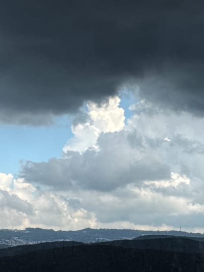 The image captures a dramatic sky over a hilly landscape, likely in or near Đa Lộc, Vietnam. The upper portion of the frame is dominated by dark, heavy storm clouds, indicative of impending rain or a passing tempest. Below these ominous clouds, patches of bright blue sky peek through, interspersed with lighter, fluffier clouds. The contrast between the dark and light clouds creates a sense of depth and atmospheric movement. The horizon is marked by a series of dark, undulating hills that recede into the distance. The foreground hills are densely covered in vegetation, appearing almost black in the low light. A scattering of buildings is faintly visible on the slopes of the middle hills, suggesting human habitation. A tall, slender structure, possibly a communication tower, is discernible on the furthest ridge. The overall impression is one of nature's power and vastness, with a hint of human presence in the rugged terrain.